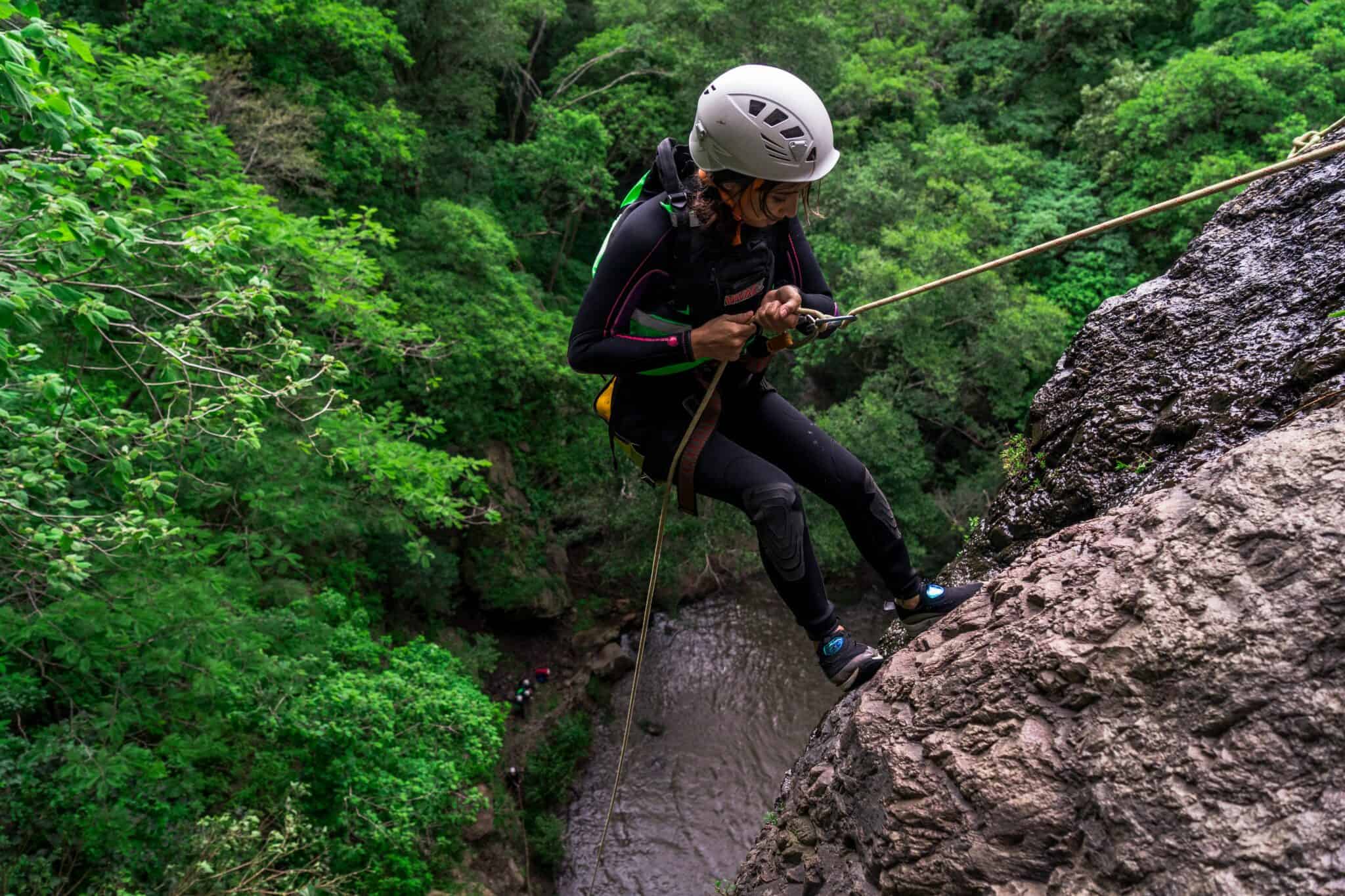 Adventurous woman rappelling down a rocky cliff in lush green forest.