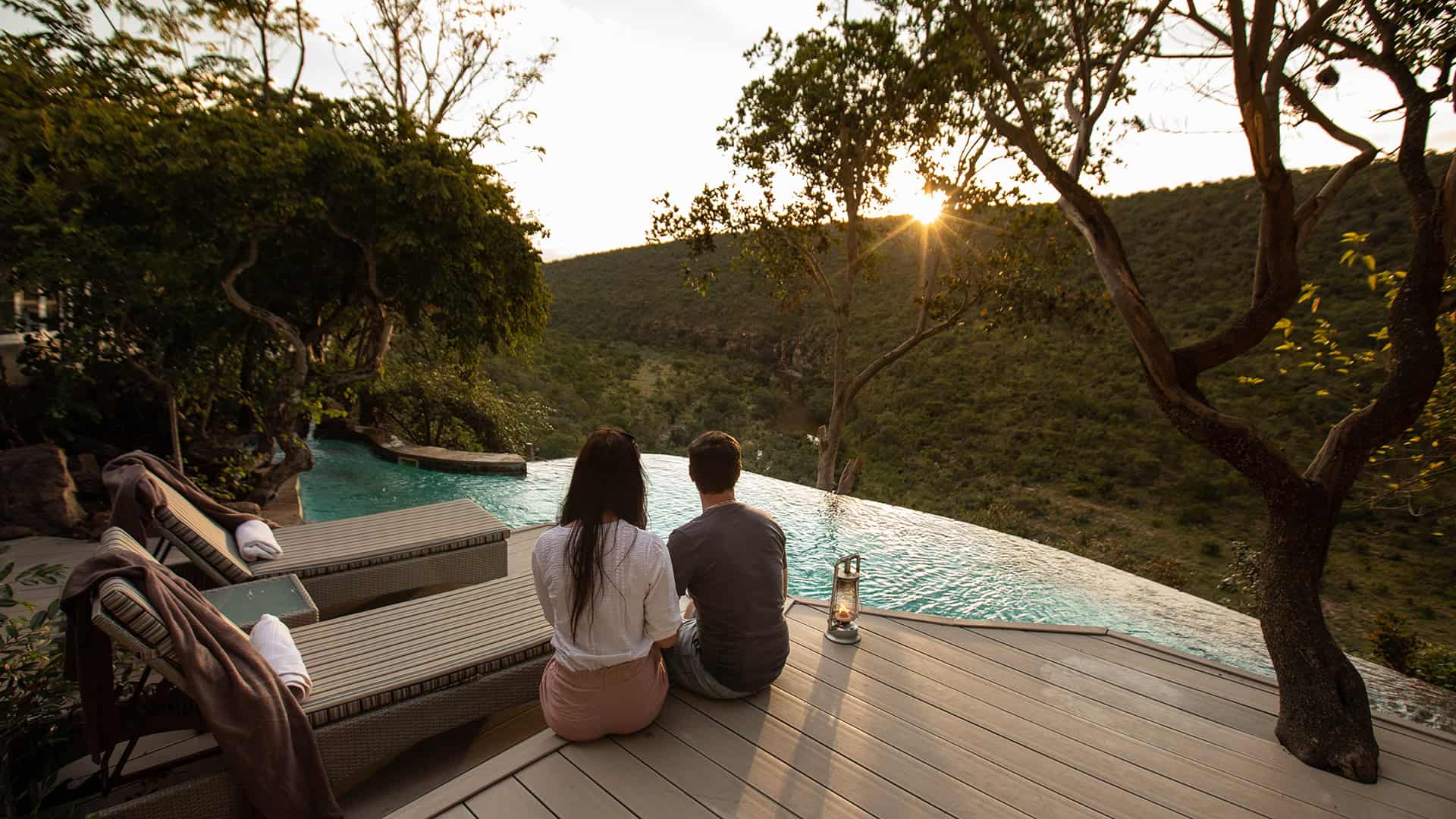Couple watching sunset over a safari landscape from a poolside deck.